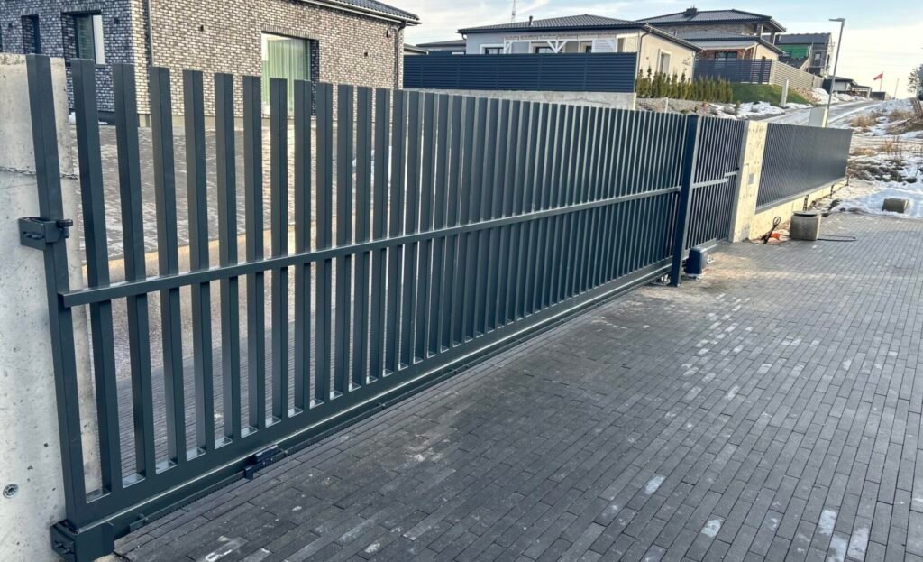 Aukšti metaliniai šukų vartai stovi šalia pastato, užtikrindami saugumą ir prieigos kontrolę.